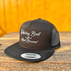 Black trucker snapback hat with 'Whiskey Bent & Hell Bound' text on a wooden surface with orange background