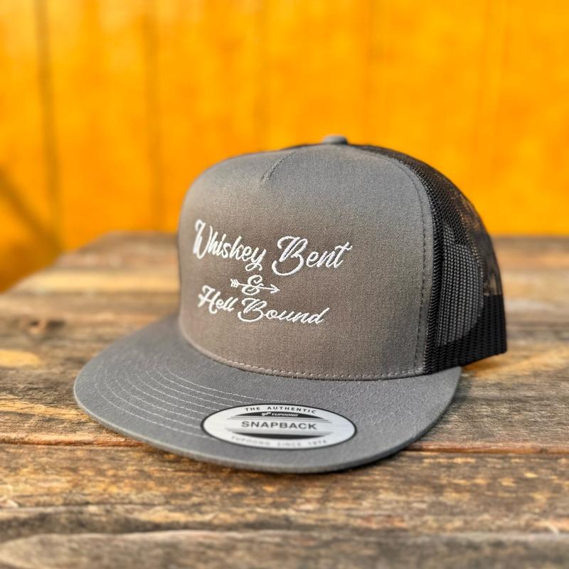 Charcoal and black trucker snapback hat with 'Whiskey Bent & Hell Bound' text on a wooden surface with an orange background.