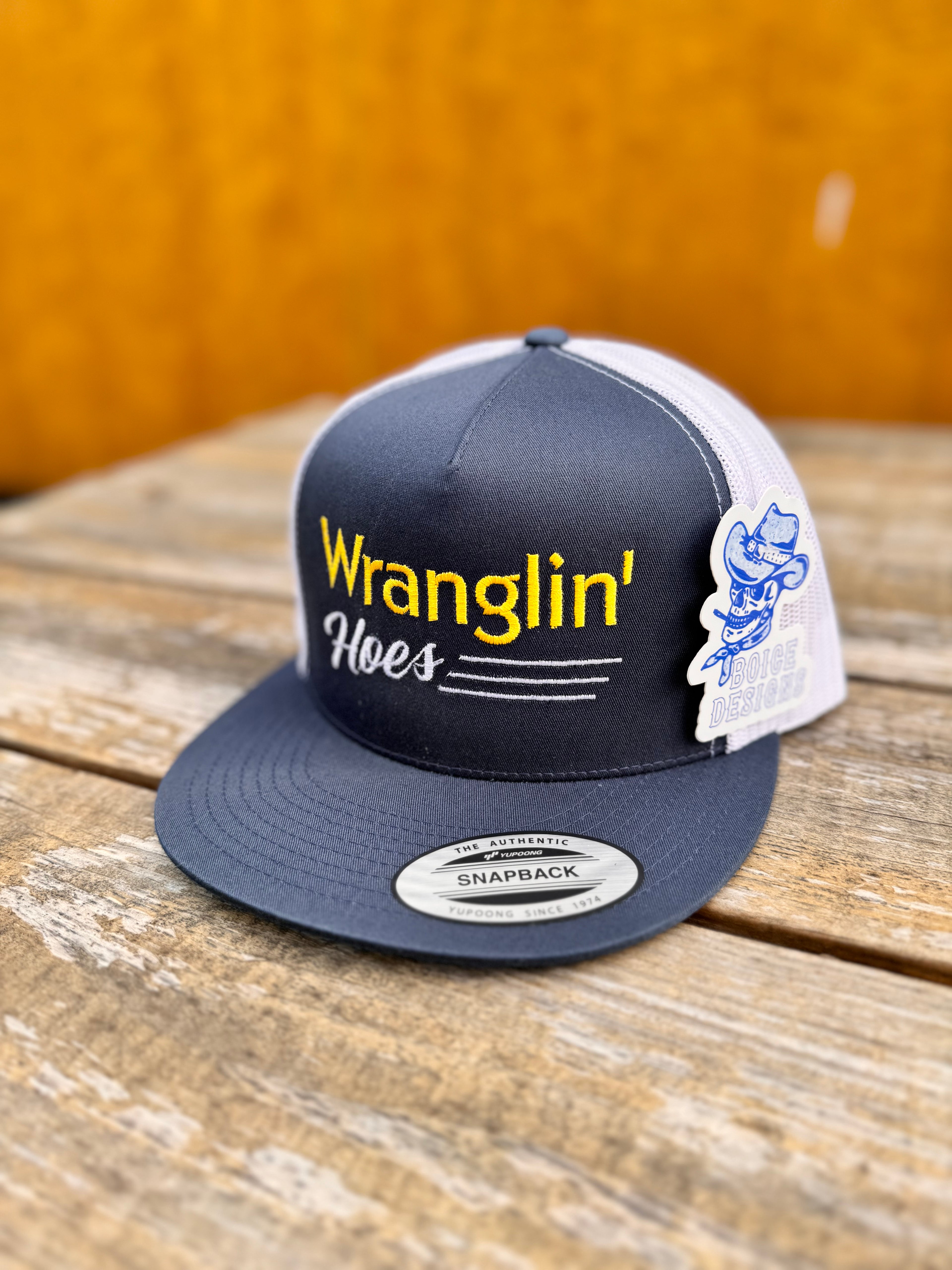 Navy and white cap with yellow and white 'Wranglin' Hoes' text on a wooden surface