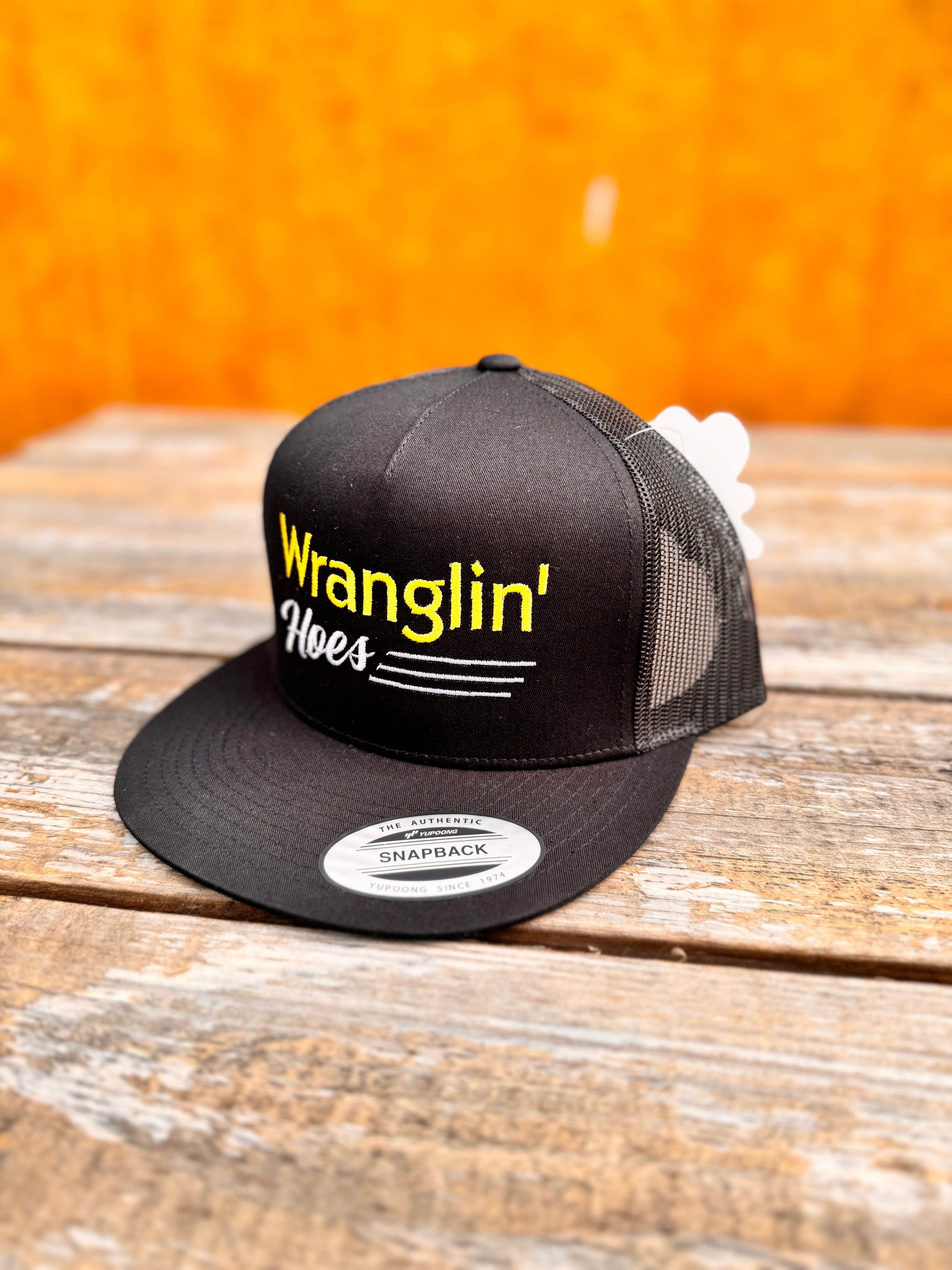 Black snapback hat with yellow and white 'Wranglin' Hoes' text on a wooden surface