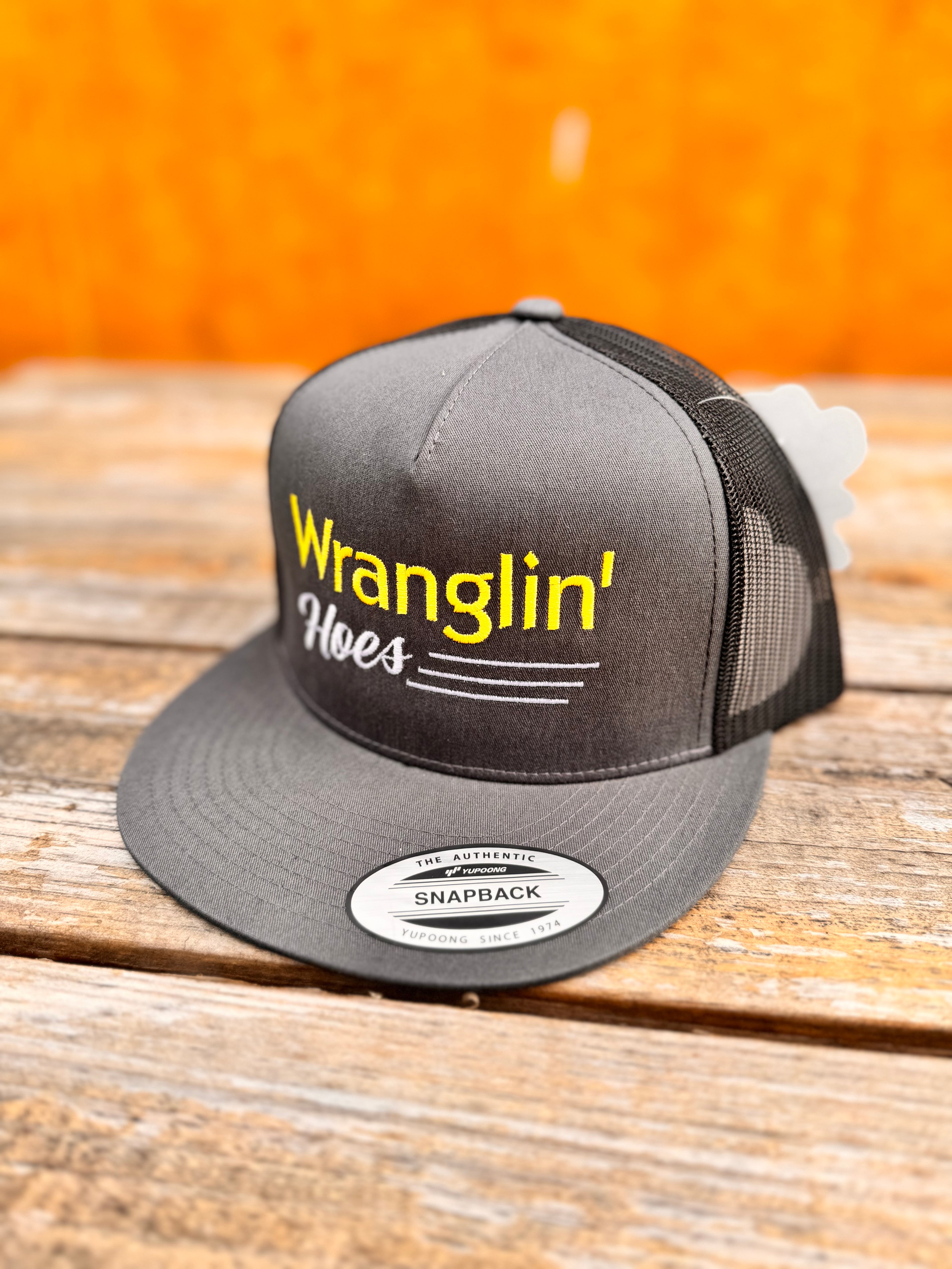 Charcoal and black snapback hat with  yellow and white 'Wranglin' Hoes' text on a wooden surface